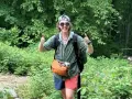 A hiker in a green, breathable hiking shirt material, gives a double thumbs-up in a lush forest.