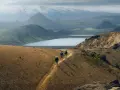 Hikers traverse a golden path on a mountain ridge, with a vast, serene lake and snow-capped peaks in the distance. This iconic landscape hints at the epic scale of how long is the Laugavegur trail.