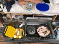 A camp kitchen checklist in action: bacon sizzles in a cast-iron pan, eggs are ready in a yellow carrier, and a portable stove is set up.