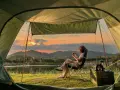 A woman enjoys solo camping by a lake at sunset, holding a camera. The tent offers a cozy view of the serene landscape.