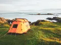 An orange tent is set up on a grassy hill overlooking a calm sea with rocky islands. This is a perfect spot for tent camp sites.