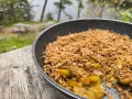 A warm, bubbling campfire apple crisp in a pan, ready to be enjoyed outdoors by a lake.