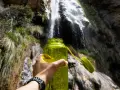 A hand holds a bright green water bottle, ready for a refreshing drink after a long hike. Behind it, a beautiful waterfall cascades down a rocky cliff, surrounded by lush greenery.