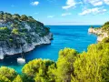 A sailboat anchors in a serene cove, surrounded by lush green trees and dramatic cliffs. This idyllic scene evokes the feeling of a perfect camping trip in Cassis.