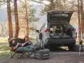 Two people enjoy a meal in camping chairs next to a car with an open trunk, showcasing clever car camping hacks with an inflatable mattress and a portable stove.