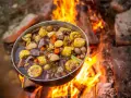 A pot of hearty camping meals, filled with corn, potatoes, carrots, and meat, simmers over a crackling campfire.