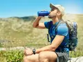 A hiker takes a refreshing drink from a blue water bottle, demonstrating how to stay hydrated while hiking in a sunny, mountainous landscape.