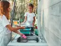 Two girls are washing dishes camping. One holds a pan over a sink while the other prepares to rinse it.
