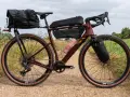 A maroon gravel bike with a full bikepacking setup, including saddle and frame bags, ready for an adventure on a dirt path.