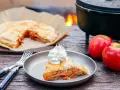 A slice of campfire apple pie, topped with whipped cream, sits on a plate next to fresh apples and a fork.