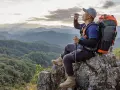 A hiker drinks water on a mountain peak, contemplating how much water to take backpacking.