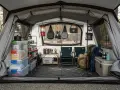 Organized camping storage inside a tent, featuring stacked bins, shelves, cooking gear, chairs, and backpacks.