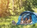 Three children laugh inside a blue tent, enjoying camping activities in a sun-dappled forest.