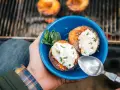 A hand holds a blue bowl with two grilled peaches topped with cream and mint. Campfire peaches are a perfect dessert.