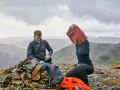 Hikers on a rocky summit adjust their backpacking clothes. One man smiles while sitting on a cairn, next to backpacks. Another person pulls on a red sweater.