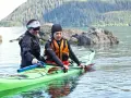 Two kayakers in a bright green kayak practice a paddle float re-entry in calm water, with a forested mountain in the background.