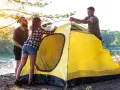 Friends setting up a bright yellow tent by a lake, learning how to choose a campsite with a beautiful sunset.