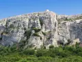 A majestic cliff face rises above a lush green forest, with a small building nestled into the rock at Sainte Baume.
