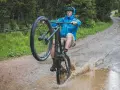 A mountain biker in a blue jacket performs a wheelie through a large puddle, splashing water as it rains heavily on a forest trail.