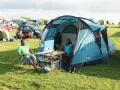 A couple enjoys a meal at their campsite, a perfect example of how to camp. They sit in chairs beside a blue tent, with food laid out on a portable table.