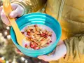 A person in a yellow jacket holds a blue bowl filled with a pink smoothie topped with granola, dried strawberries, and banana slices. Perfect for camping breakfast ideas.
