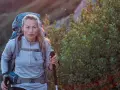 A woman with blonde hair, wearing a grey jacket and a large backpack, is backpacking hiking up a rocky, vegetated slope. She holds a trekking pole.