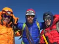 Three smiling mountaineers in bright down fill jackets and helmets pose on a sunny summit, ready for adventure.