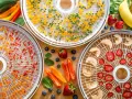 Dehydrating food for backpacking: trays filled with dried peppers, corn, apples, and strawberries, surrounded by fresh produce.