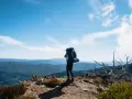 A lone hiker stands on a mountain ridge, ready for backpacking and hiking, with a vast expanse of blue mountains stretching to the horizon under a clear sky.