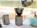 Making coffee while camping: hot water pours from a kettle into a pour-over filter, brewing coffee into a metal mug. A bag of Sisters Coffee Guatemala sits nearby.