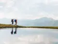 Two hikers with backpacks walk along a grassy ridge beside a calm lake, with misty mountains in the background. This image inspires thoughts on how to go backpacking.