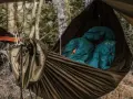 Cozy hammock camping setup in a forest. Two teal sleeping bags are nestled inside a brown hammock, ready for a night under the stars.