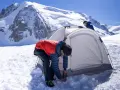 Two people set up a tent for winter camping amidst a snowy mountain landscape under a clear blue sky.