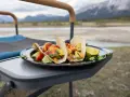 Delicious camping tacos with avocado, tomatoes, and lime, served on a plate on a portable table with mountains in the background.