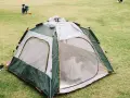 A green and gray tent is partially set up on a grassy field. A child plays in the background, learning how to set up a tent.