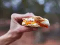 A hand holds a gooey white chocolate s'more, its toasted marshmallow and jam oozing out.