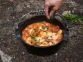 A hand garnishes a hearty dutch oven paella with fresh parsley. This campfire meal features shrimp, sausage, and rice in a rich tomato sauce.