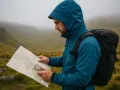 A hiker in a blue jacket uses a compass on a map to navigate a foggy mountain landscape.