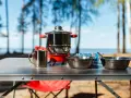 A complete camping kitchen setup with a stove, pots, bowls, and mugs on a table, ready for outdoor cooking by a lake.