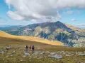 Hikers with backpacks traverse a rocky, grassy meadow on a pyrenees hut to hut hiking adventure, with majestic mountains under a cloudy sky.