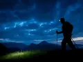 Silhouette of a hiker with a headlamp on, trekking poles in hand, on a night hike through a dramatic, cloudy sky over mountains.