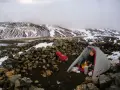A hiker prepares a meal inside a tent at one of the Laugavegur trail huts, surrounded by snow-dusted mountains and a stone wall.