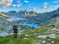 Hikers ascend a grassy slope on the HRP trail, with a stunning mountain lake and jagged peaks in the background under a cloudy sky.