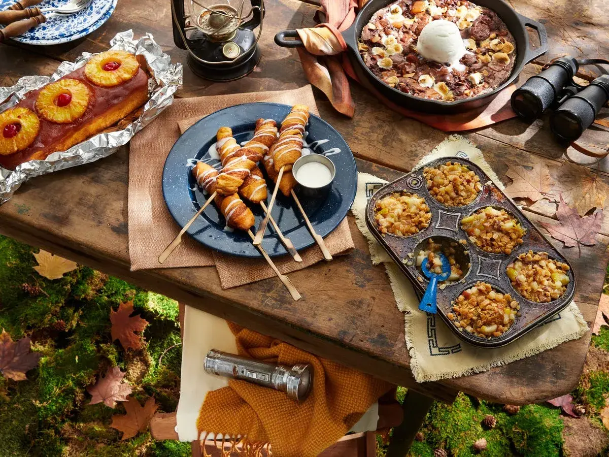 A spread of cozy camping food: pineapple upside-down cake, pigs in a blanket, apple crumble muffins, and a skillet brownie with ice cream.