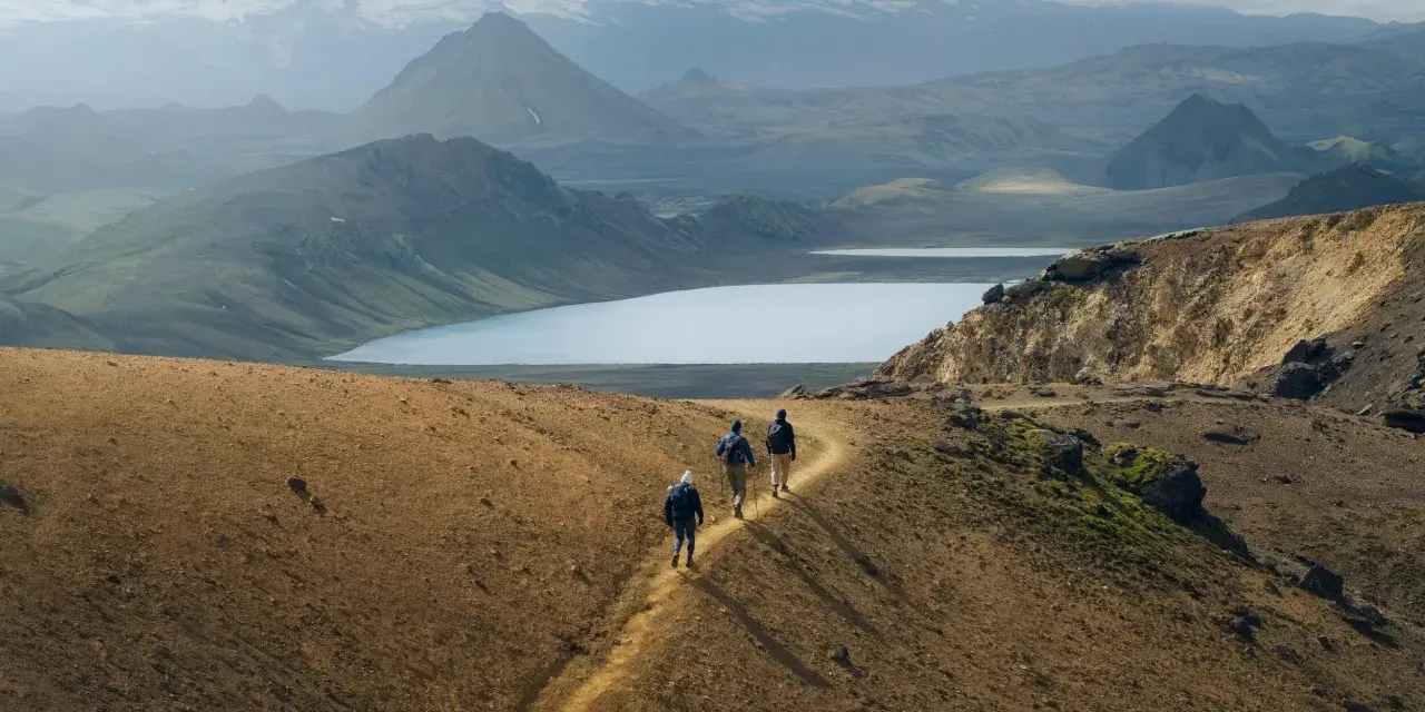 Hikers traverse a golden path on a mountain ridge, with a vast, serene lake and snow-capped peaks in the distance. This iconic landscape hints at the epic scale of how long is the Laugavegur trail.