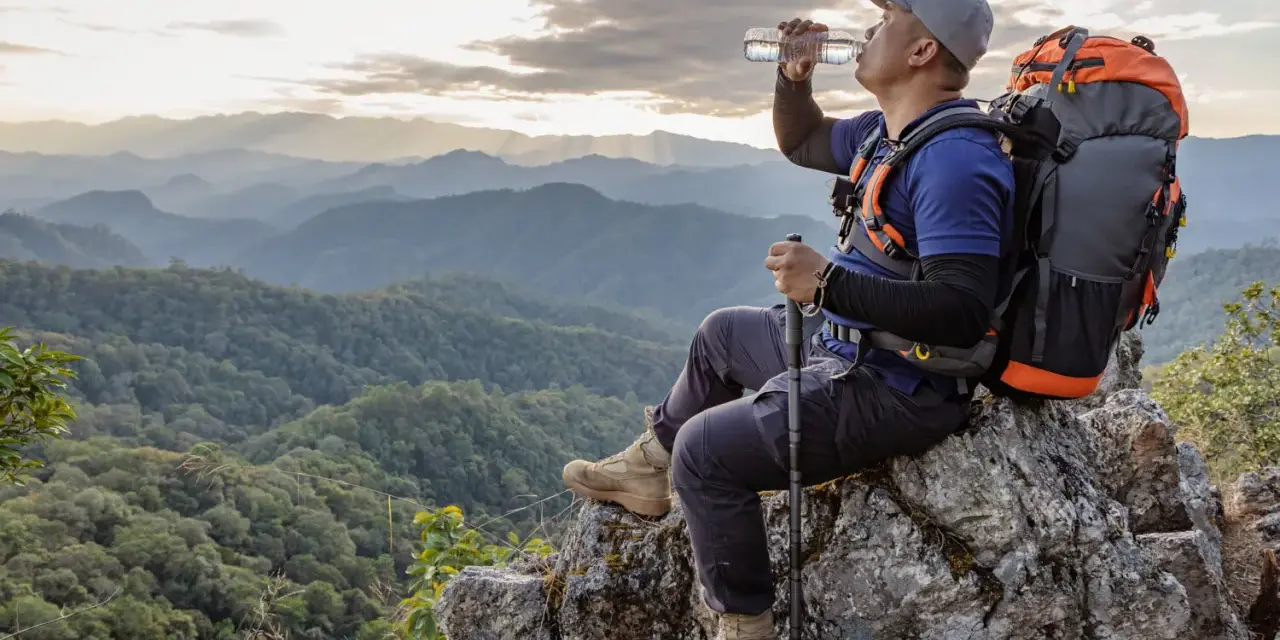 Backpacker drinks water on a rocky outcrop, contemplating how much water to bring backpacking for this stunning mountain vista.