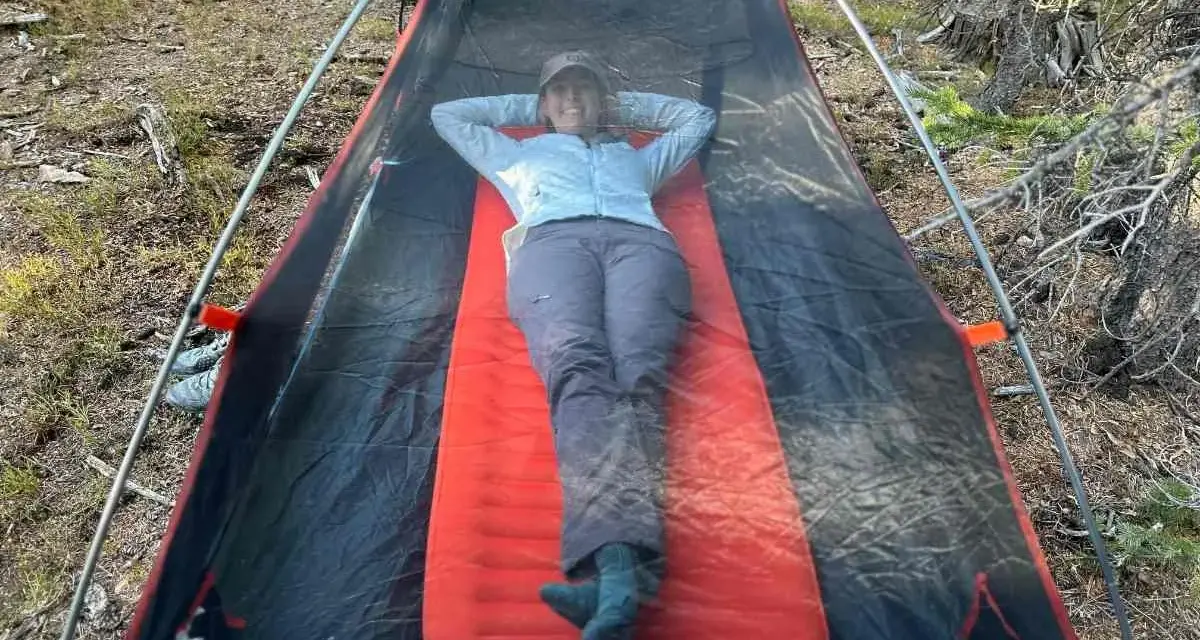 A person relaxes inside a tent on a bright orange Therm-a-Rest NeoAir XLite sleeping pad, enjoying the outdoors.