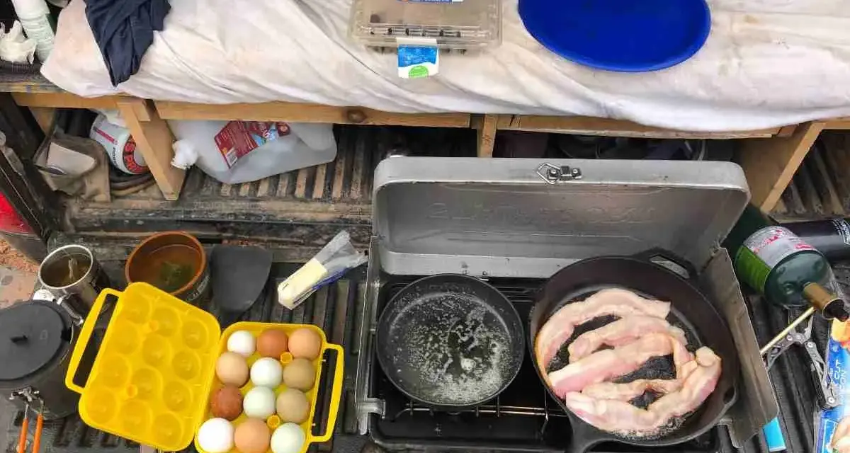 A camp kitchen checklist in action: bacon sizzles in a cast-iron pan, eggs are ready in a yellow carrier, and a portable stove is set up.