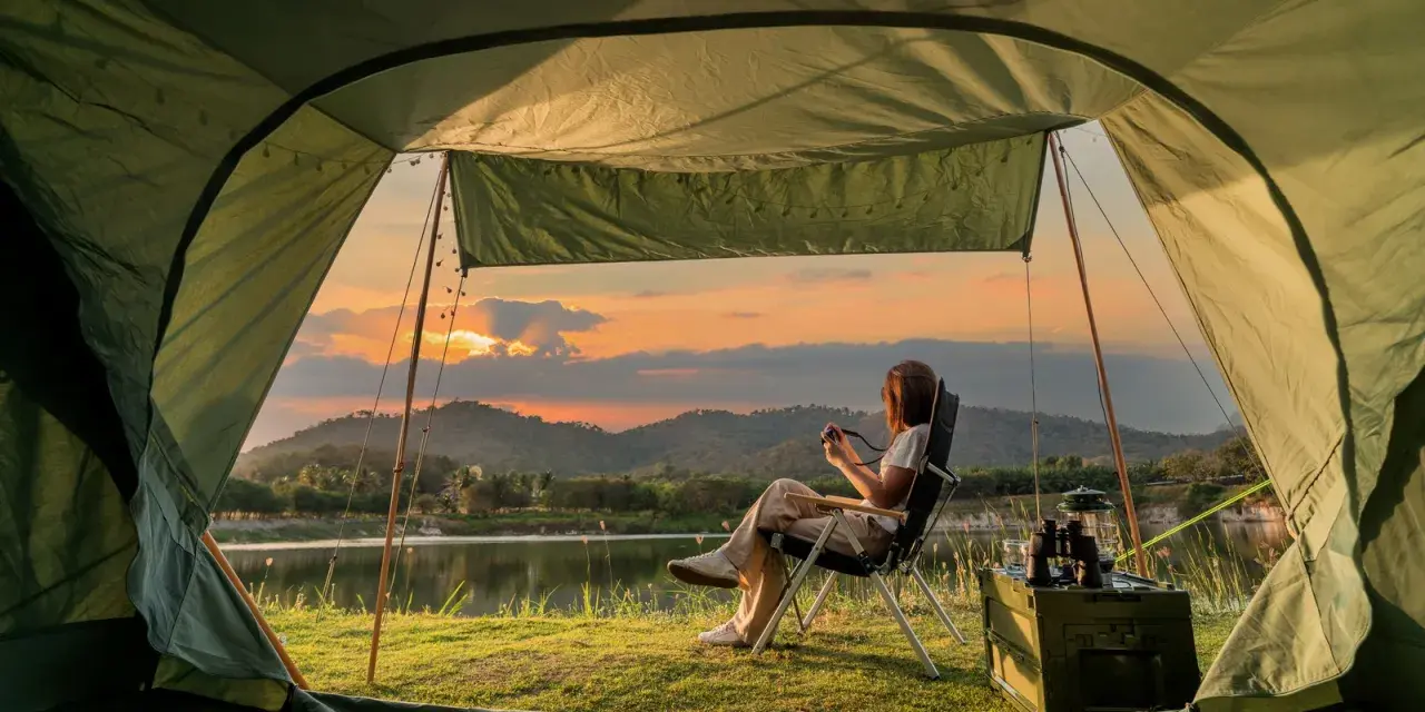 A woman enjoys solo camping by a lake at sunset, holding a camera. The tent offers a cozy view of the serene landscape.
