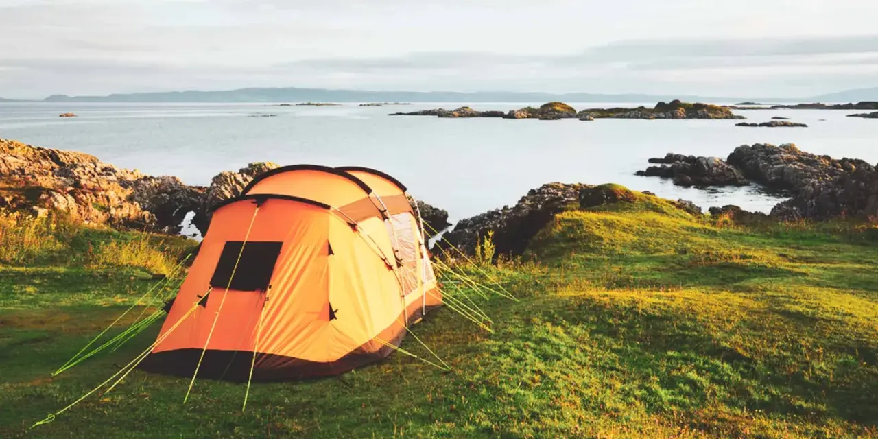 An orange tent is set up on a grassy hill overlooking a calm sea with rocky islands. This is a perfect spot for tent camp sites.